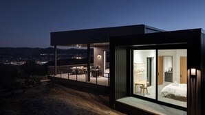 A modern house with a glass-enclosed bedroom, a balcony with outdoor seating, and a city view at dusk.
