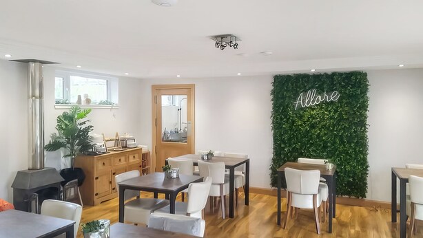 Un área de comedor moderna con mesas y sillas de madera, una pared verde con un letrero y un mueble de madera con utensilios de cocina.