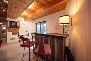 A cozy room with wooden ceiling beams, a dartboard, a moose-shaped chair, and a lamp.