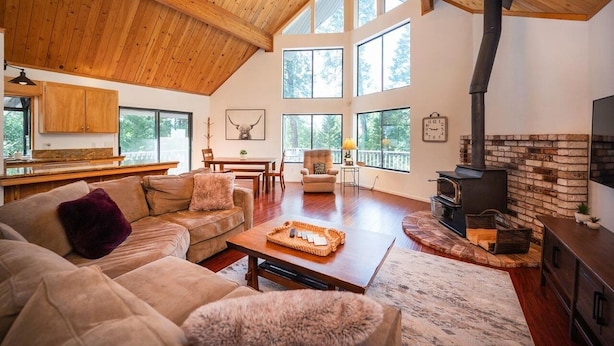 Amplia sala de estar con techo alto, vigas de madera, chimenea y grandes ventanas que ofrecen vistas al exterior.