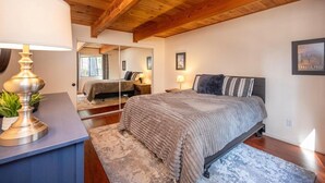 Un dormitorio con techo de madera, una cama con manta gris, una mesita de noche con lámpara y un espejo que refleja la habitación.