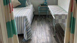 Une chambre à coucher avec un lit, une petite table, une fenêtre avec des stores et une œuvre d’art représentant un hippocampe accrochée au mur.