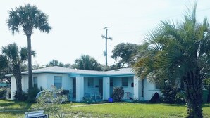 Une maison de plain-pied avec un toit bleu, entourée de palmiers et d’une pelouse verdoyante.