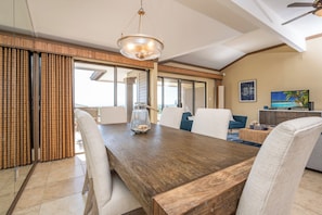 Un vaste espace dédié à la salle à manger, avec une table en bois, des chaises blanches, un lustre et une vue sur l’extérieur.