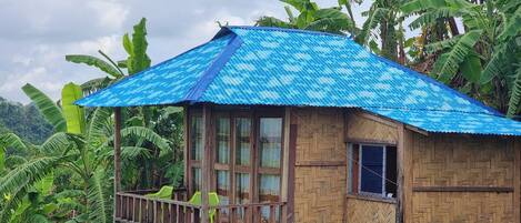 Una casa de madera con techo de metal corrugado azul, rodeada de exuberantes plantas de banano.