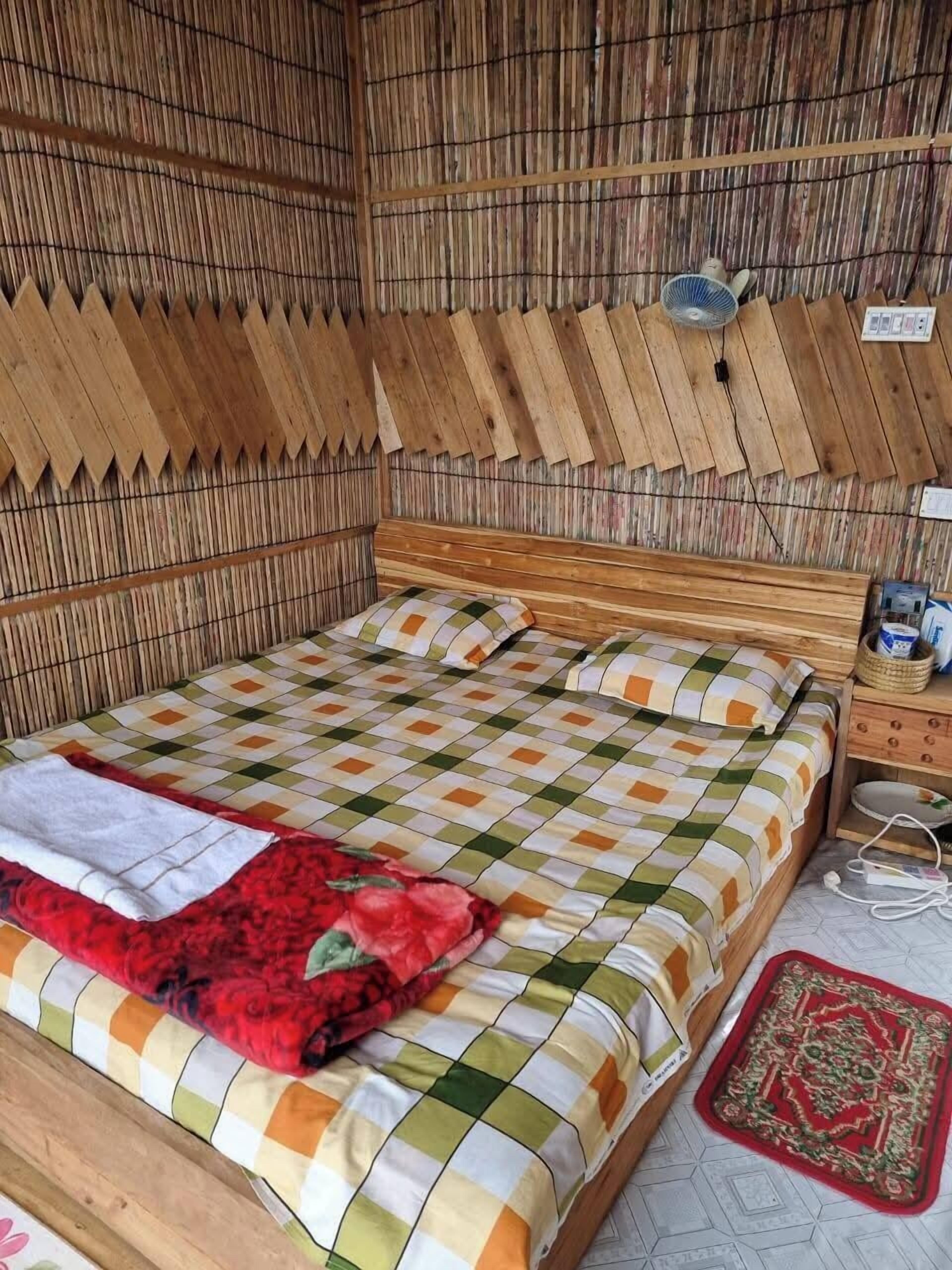 A room with a wooden bed, a checkered bedspread, a red blanket, and a wooden nightstand with a lamp.