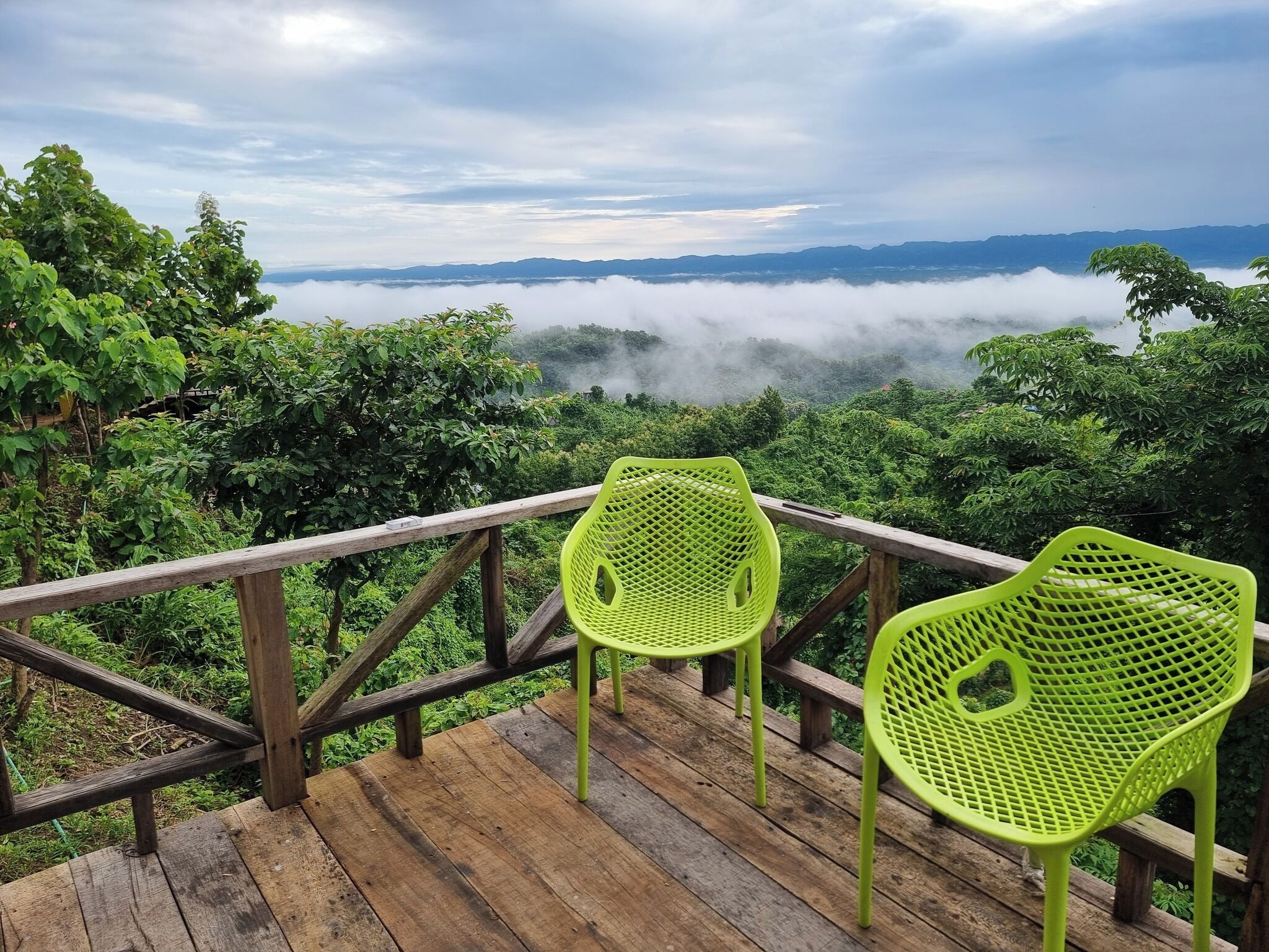 A wooden deck with two bright green chairs overlooking a lush landscape with mountains and a foggy valley.