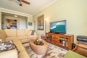 A living room with a sofa, television, wooden cabinet, and a wicker coffee table. - Kapalua Golf | Golf View 6 BR, Sleeps 18 | Car Incl. w/ 6+ Nights | KGV ML-2327 by KBM (Lahaina)
