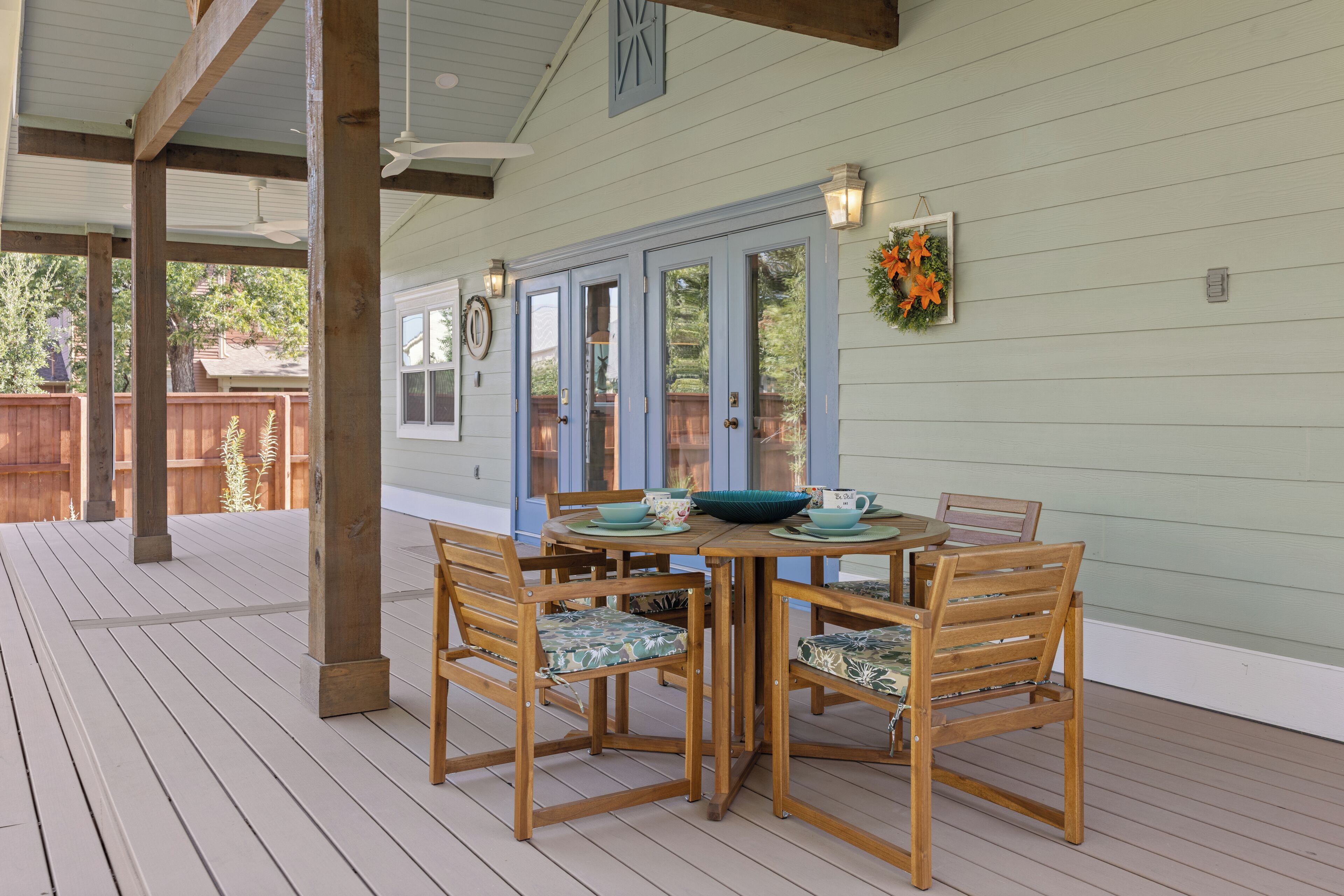 A covered outdoor dining area with a table and chairs, wooden beams, and a view of a house through glass doors.
