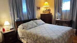 Un lit bien fait, recouvert d’une couverture matelassée, une table de chevet avec une lampe et une horloge, une commode et un bureau avec une lampe et un ventilateur.