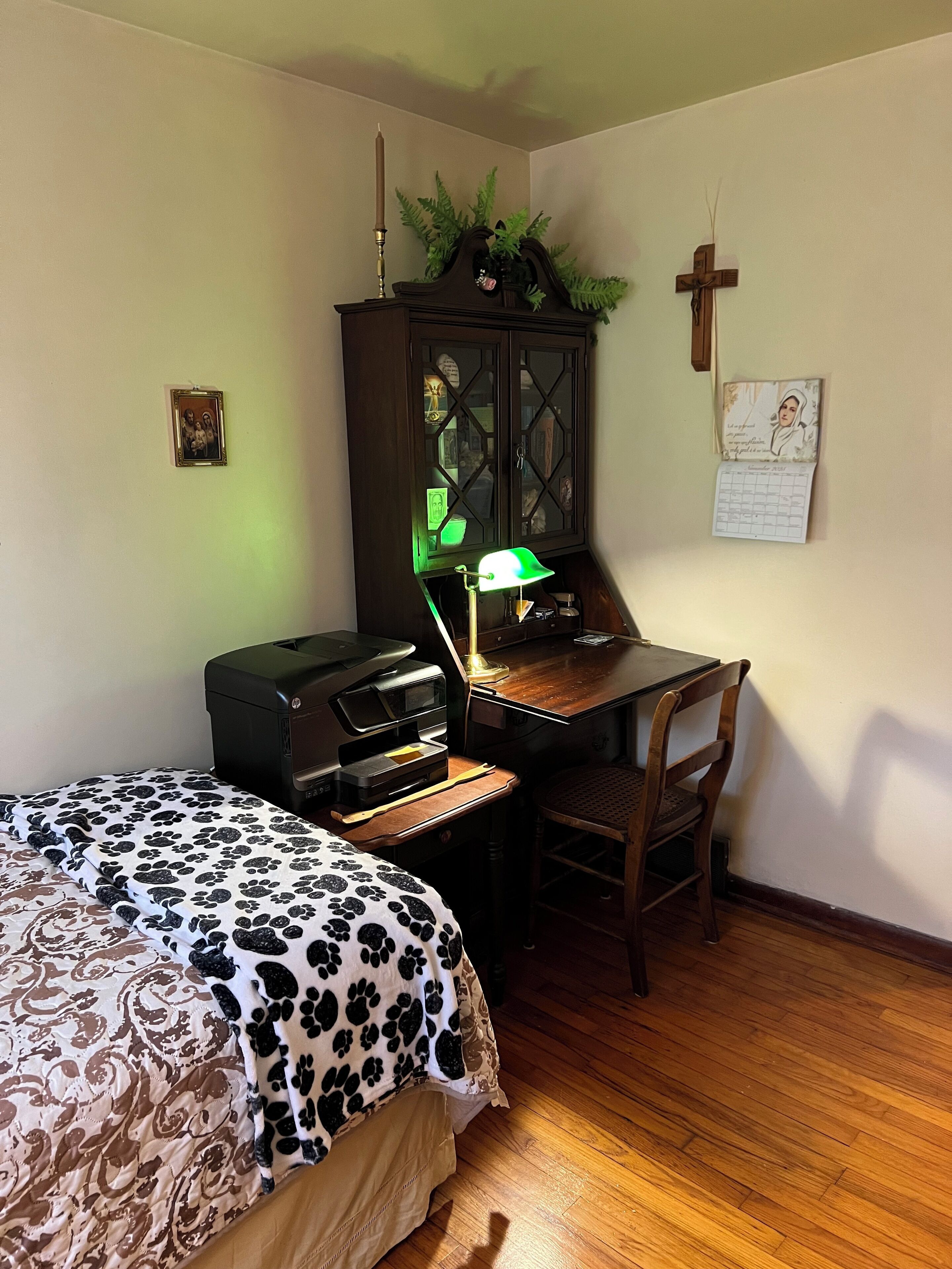 A bedroom with a bed, a desk with a lamp, a printer, and a cross on the wall.