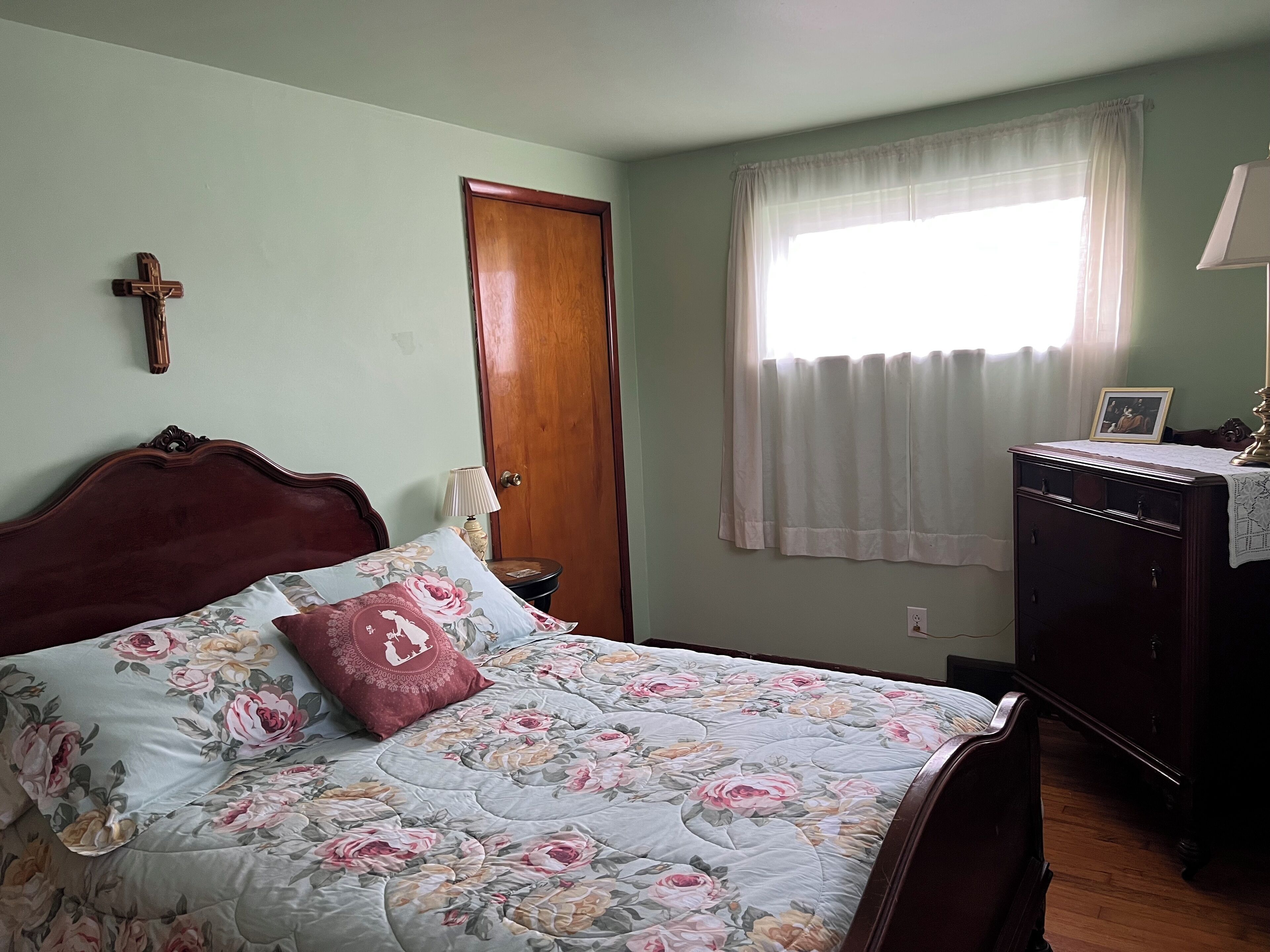 Ein Schlafzimmer mit einem Holzbett, floralem Bettbezug, einem Holzschrank, einer Nachttischlampe, einem hölzernen Kreuz an der Wand und einem Fenster mit Vorhängen.