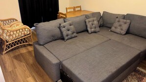 A grey sofa with four cushions, a wicker chair, a wooden dining table, and a black curtain.