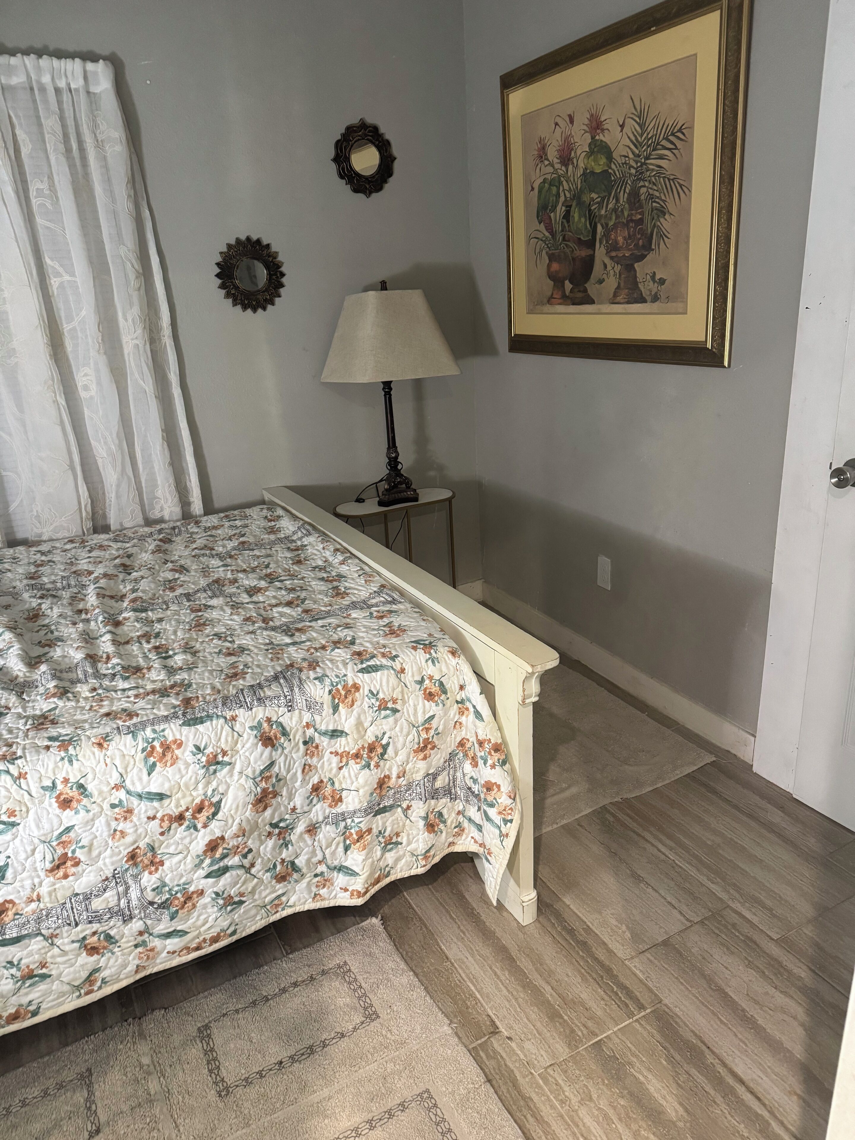 A bedroom with a bed, a nightstand, a lamp, a mirror, and a framed botanical painting on the wall.