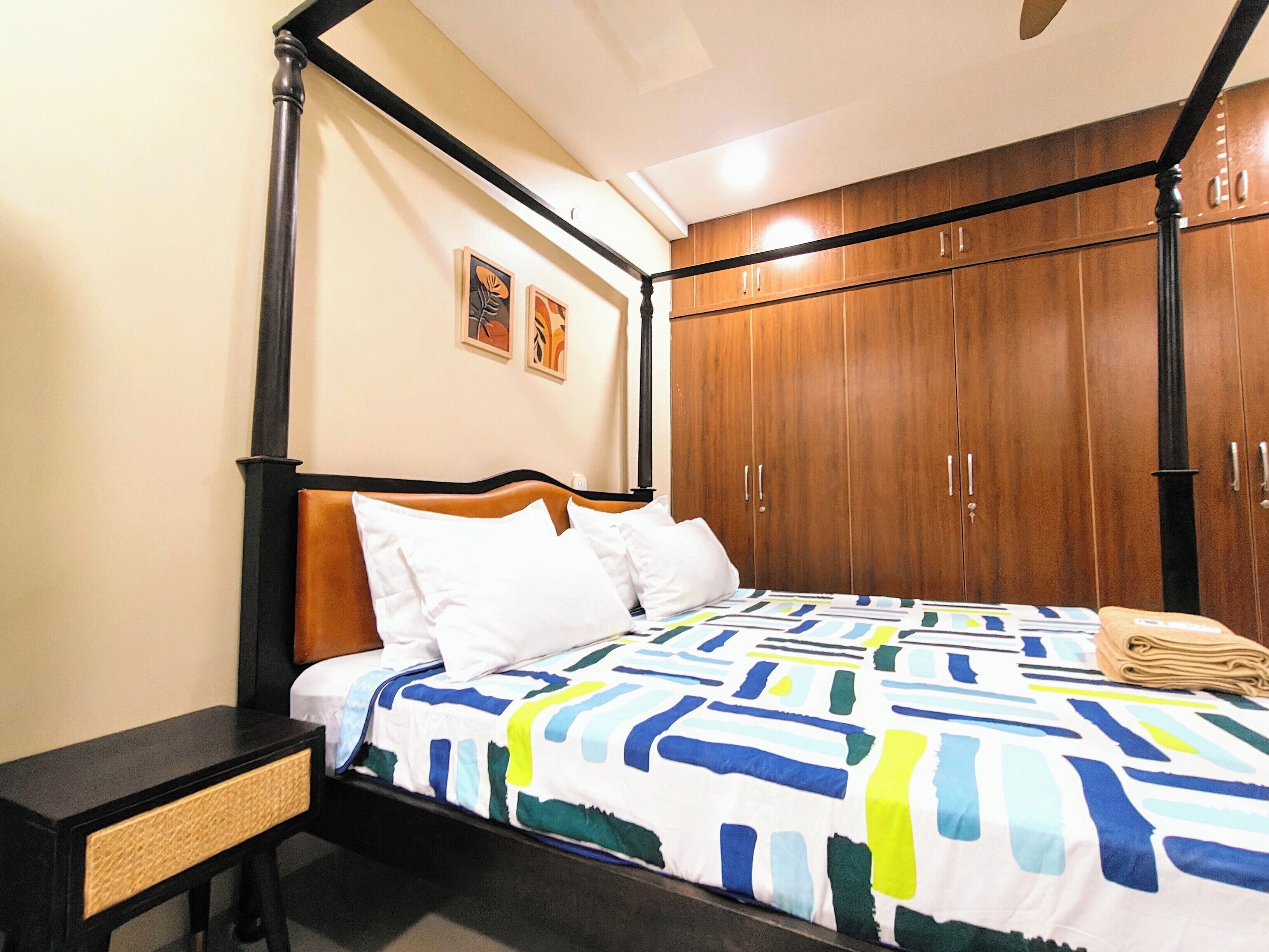 A four-poster bed with a colorful geometric-patterned bedspread, a wooden wardrobe with multiple doors, and a small bedside table with a woven drawer.