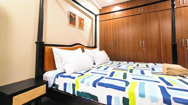 A four-poster bed with a colorful geometric-patterned bedspread, a wooden wardrobe with multiple doors, and a small bedside table with a woven drawer. - Crescent Stays Banjara Hills  (Hyderabad)