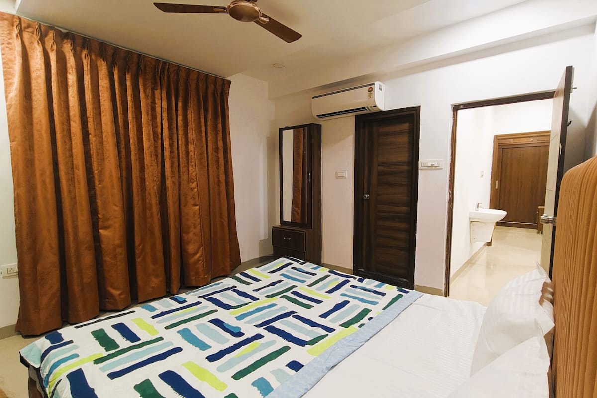 A bedroom with a bed, a ceiling fan, an air conditioning unit, a wardrobe, and a bathroom visible through an open door.