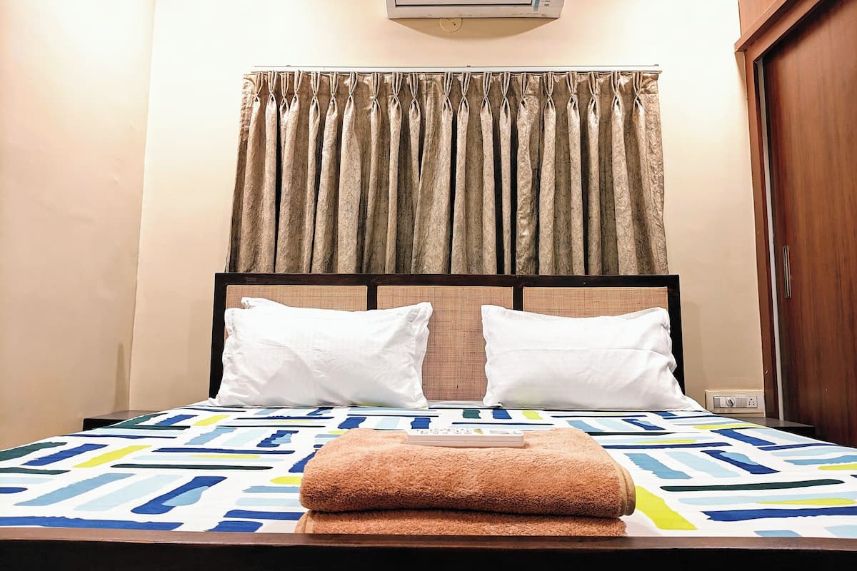 A hotel room with a bed, two pillows, a blanket, a remote control, and an air conditioning unit on the wall.
