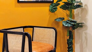 Una obra de arte enmarco sobre una pared amarilla, una silla de mimbre con un cojín marrón y una planta verde alta.
