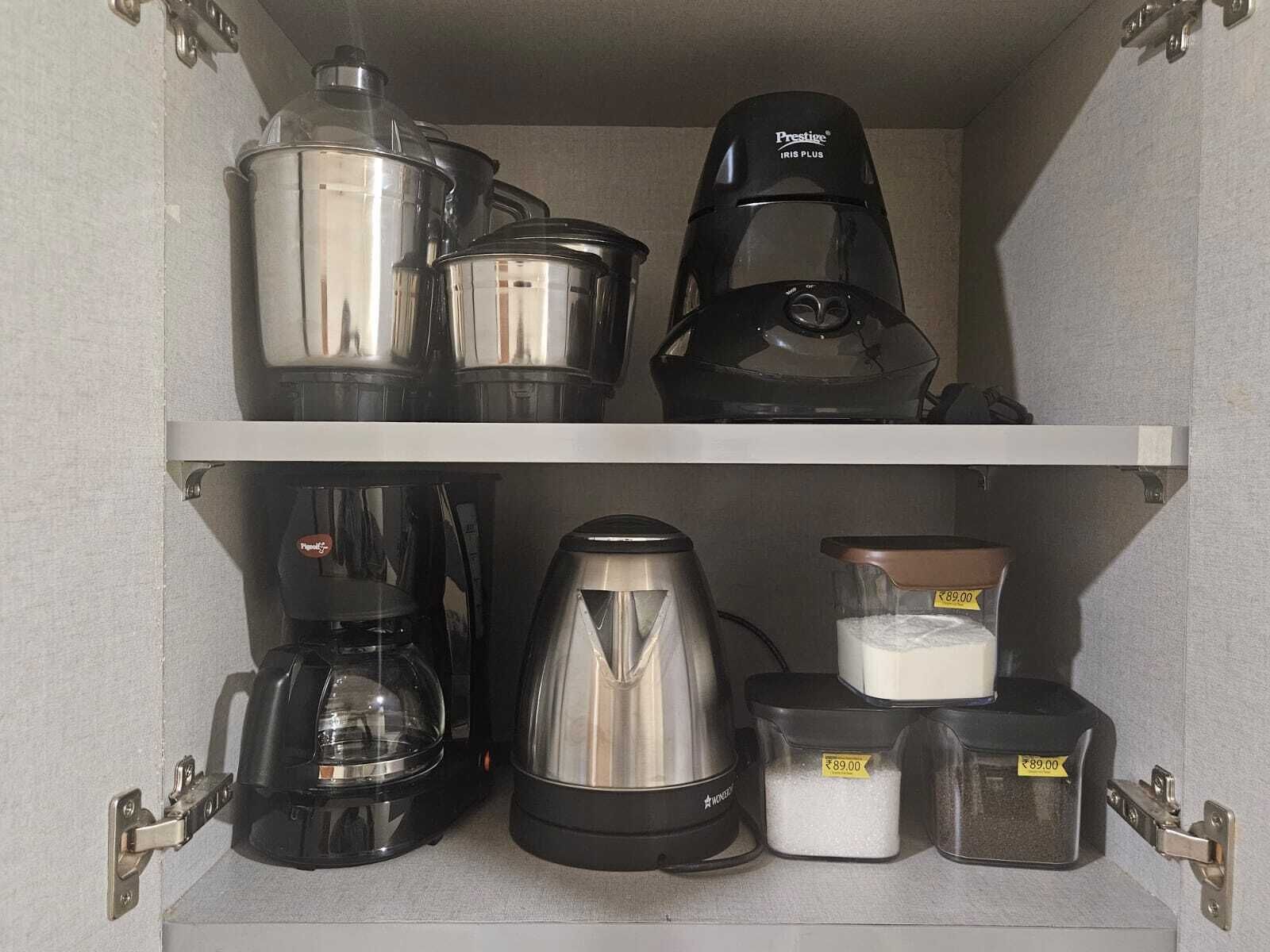 A kitchen cabinet with various appliances including a coffee maker, a kettle, and containers with white powder.