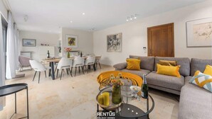 Een moderne woonkamer met een grijze bank, gele kussens, een eethoek met witte stoelen, en een keuken op de achtergrond.