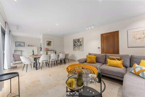 A modern living room with a grey sofa, yellow cushions, a dining area with white chairs, and a kitchen in the background. - Villa Riposo - charming villa by the sea Koksijde (Koksijde)
