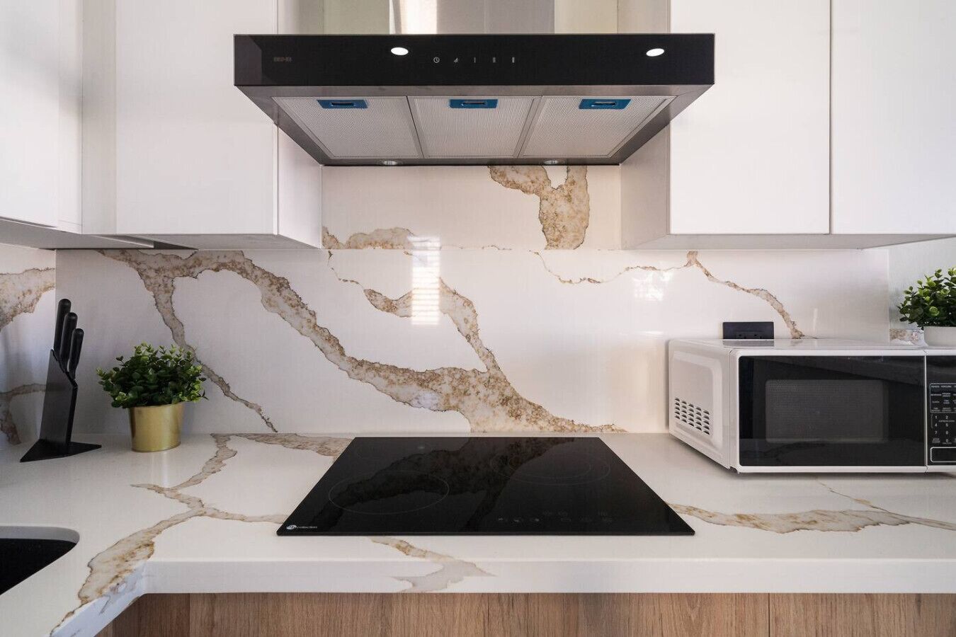 A modern kitchen with a marble backsplash, a black induction cooktop, a white microwave, and a black range hood.