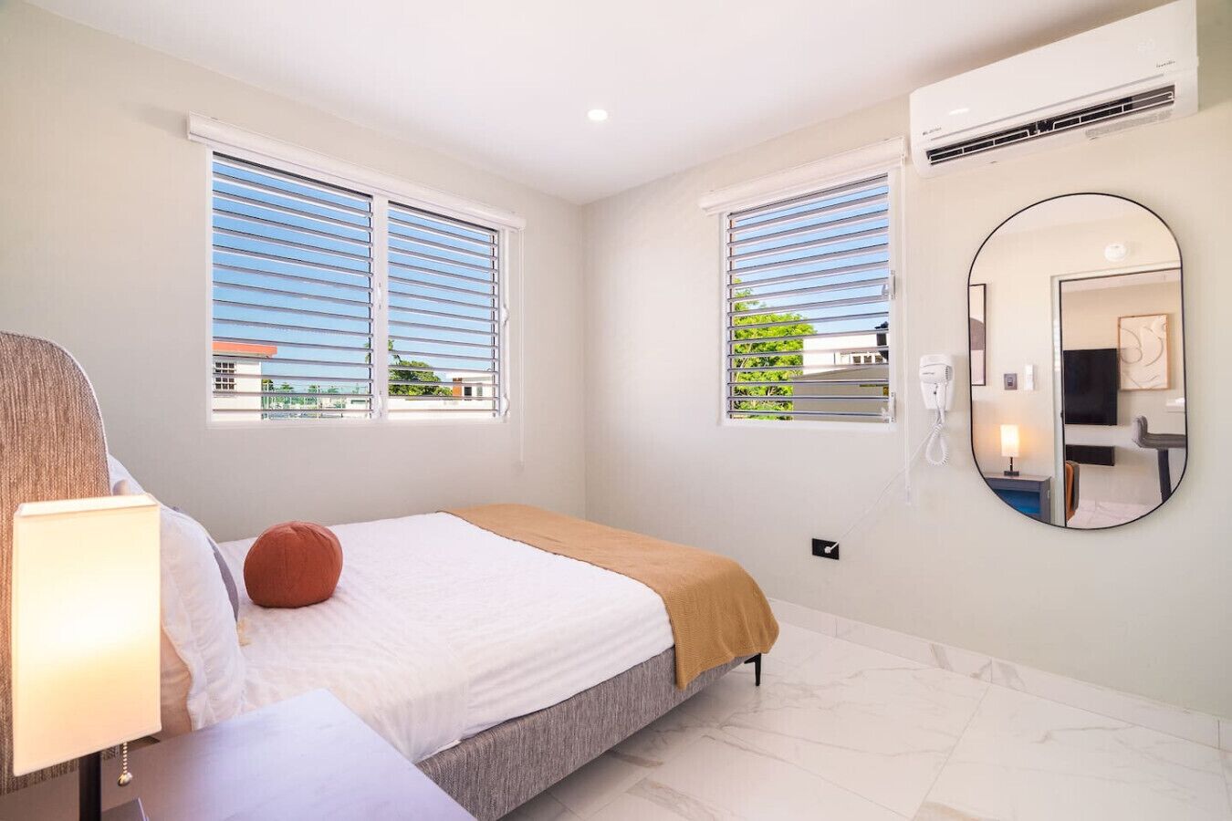 Habitación de hotel con una cama, una mesita de noche con lámpara, un espejo redondo y dos ventanas con persianas.