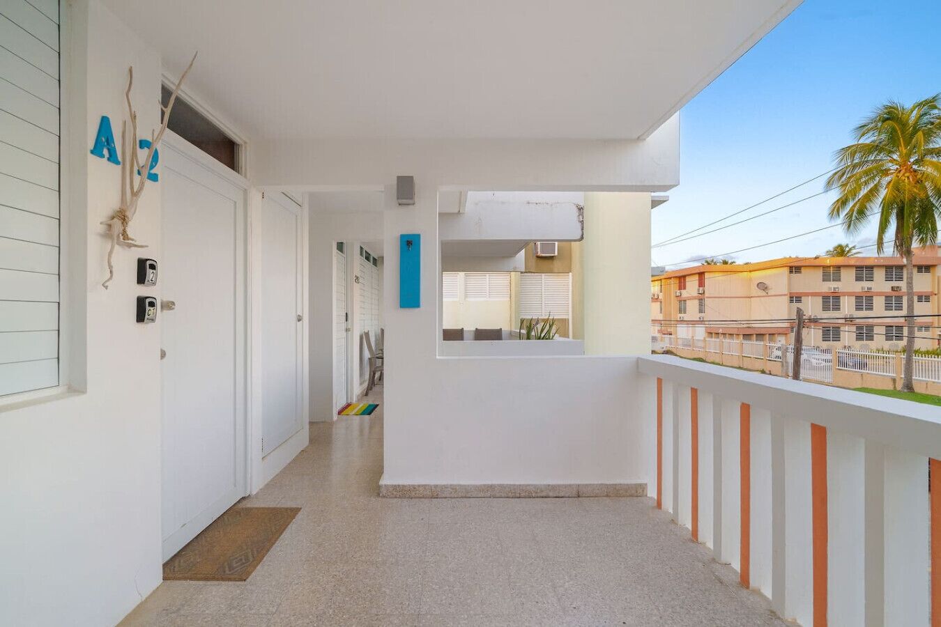 Un balcon donnant sur des palmiers et des immeubles, une porte blanche avec un numéro, et une petite fenêtre.