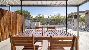 Um pátio externo com mesa e cadeiras de madeira, cerca de metal e área coberta com telhado metálico.