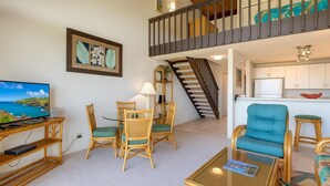 A living room with a TV, dining table, chairs, a sofa, a staircase, and a kitchen area. - Napili Point | Oceanfront 3 BR, Sleeps 10 | Car Incl. w/ 6+ Nights | NAP ML-2438 by KBM (Lahaina)