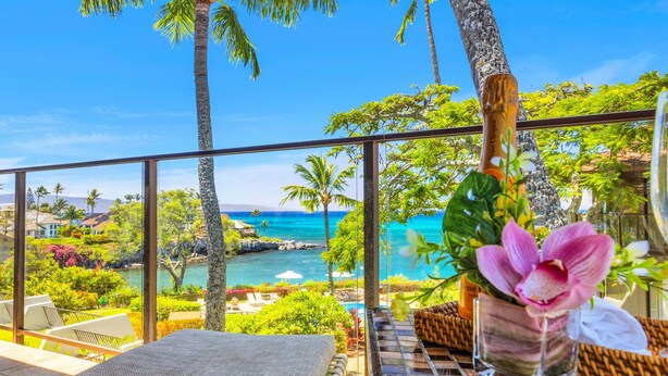 Un balcon donnant sur une plage et l’océan, une chaise en osier et un vase rempli de fleurs.