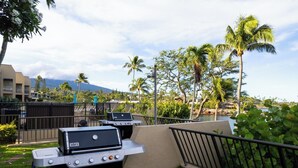 Eine Terrasse mit zwei Grillstationen, einem Tisch und einem Geländer mit Blick auf eine angelegte Grünfläche, Palmen und eine Bergsicht.