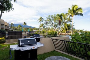 A patio with two barbecue grills, a table, and a railing overlooking a landscaped area with palm trees and a view of a mountain. - Napili Point | Oceanfront 7 BR, Sleeps 20 | Car Incl. w/ 6+ Nights | NAP ML-2444 by KBM (Lahaina)