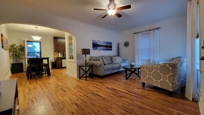 A living room with a sofa, a coffee table, and a dining area with a table and chairs. There is a ceiling fan, a window with curtains, and a painting on the wall. - Clean quiet and comfortable home welcomes you (Colorado Springs)