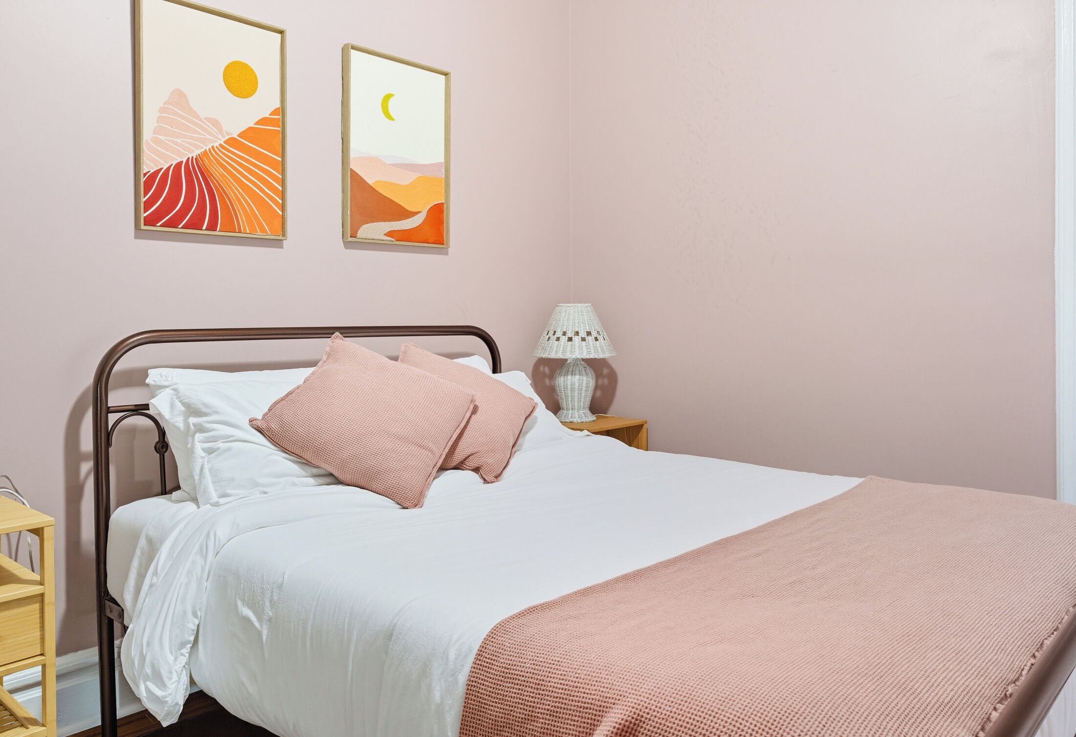 Ein ordentlich bezogenes Bett mit einer weißen und rosa Decke, einer Nachttischlampe und zwei gerahmten Bildern an der Wand.