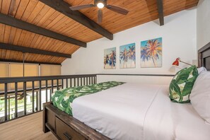 A bedroom with a wooden ceiling, a bed with a green leaf-patterned bedspread, a red desk lamp, and three palm tree paintings on the wall. - Napili Point | Oceanfront 3 BR, Sleeps 8 | Car Incl. w/ 6+ Nights | NAP ML-2456 by KBM (Lahaina)