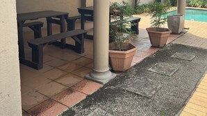 Ett takförsett utomhusområde med sittplatser och bänkar, samt krukväxter, en pool i bakgrunden och ett klinkergolv.