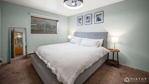 Een slaapkamer met een bed, een nachtkastje, een lamp, een plafondlamp, een raam met zonwering en ingelijste foto's aan de muur.