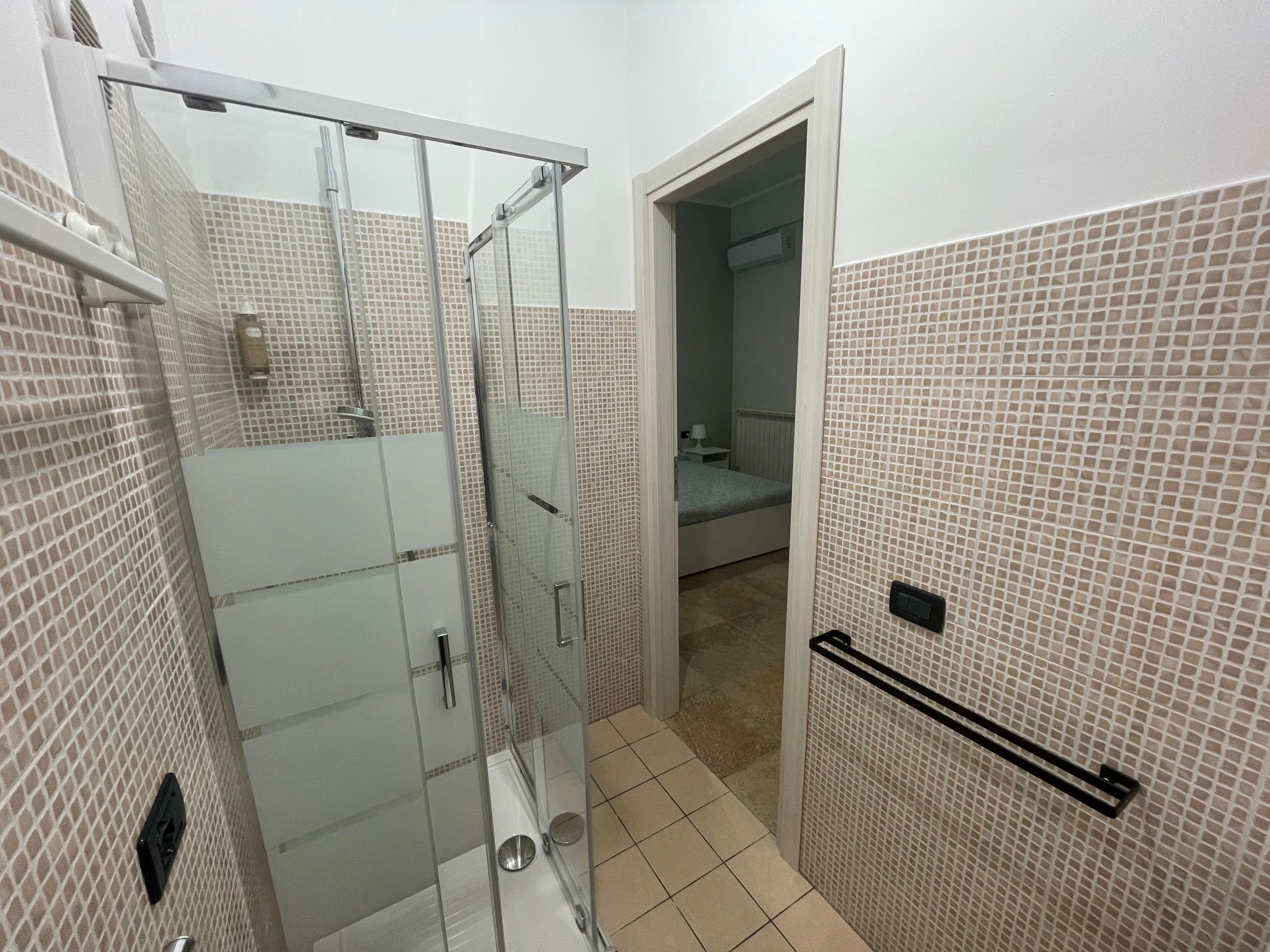 Een badkamer met een glazen douchecabine, een betegelde vloer en een bed dat door een geopende deur zichtbaar is.