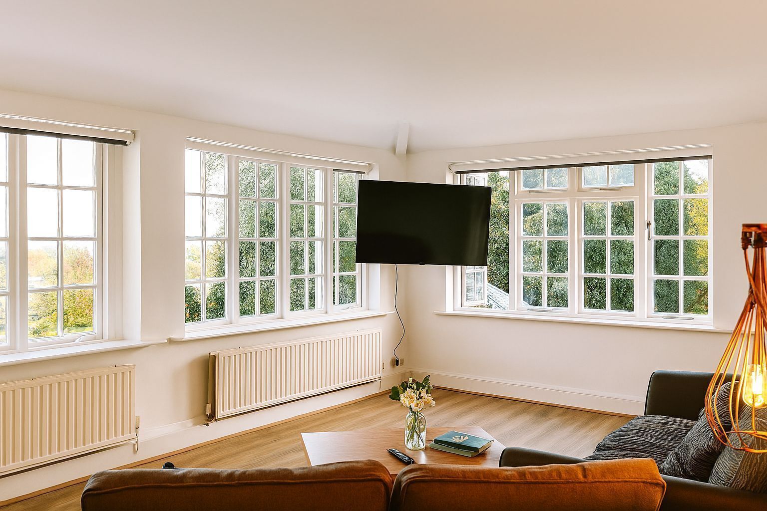 Ein geräumiges Wohnzimmer mit großen Fenstern, einem Flachbildfernseher, Holzboden, einem braunen Sofa, einem kleinen Tisch mit einer Vase und einer außergewöhnlichen Pendelleuchte.