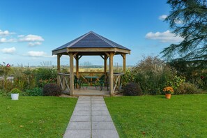 Ein hölzerner Gazebo mit einem Ziegeldach, umgeben von einem gepflegten Garten und wolkenlosem Himmel.