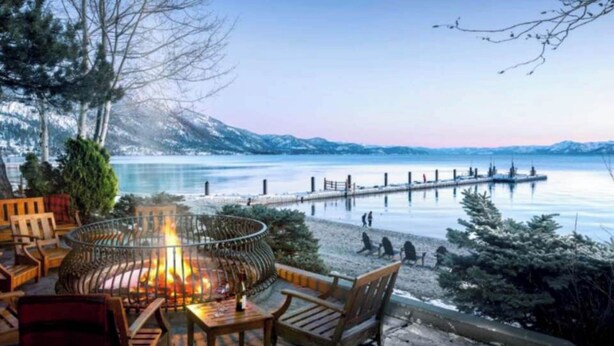 Un espace de détente aménagé au bord du lac, avec un foyer, des meubles en bois et une vue sur une jetée qui s’avance dans l’eau.
