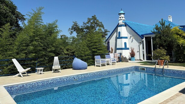 Piscina con sillas de descanso y una pequeña mesa, rodeada de vegetación y un edificio con techo azul y paredes blancas.