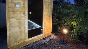 Una cabaña de madera con un amplio ventanal, suelo de grava y una pequeña luz exterior.