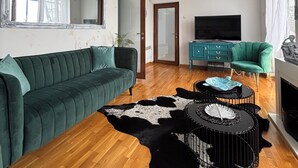 Una sala de estar con un sofá verde, suelo de madera, alfombra de vaca en blanco y negro, un mueble azul aguamarina y un televisor.