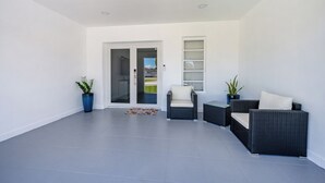 En modern uteplats med en mörkrottingsoffa, en vit sittstol med vaddering och två krukväxter.