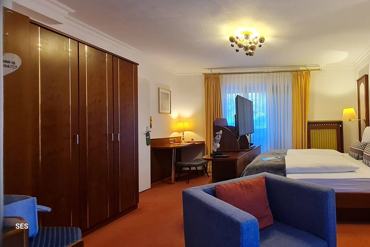 A hotel room with a double bed, a desk with a lamp, a chair, a wardrobe, and a television.