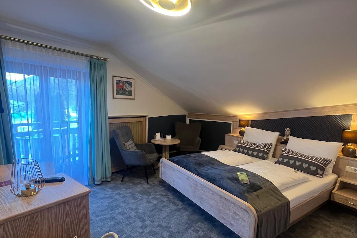 A modern hotel room with a large bed, a desk, and a chair. There is a window with curtains and a view of greenery outside.