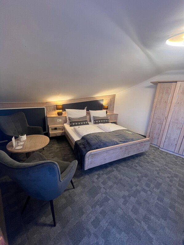 A bedroom with a bed, two bedside tables, a chair, a small table, and a wardrobe. - Hotel Quellenhof (Bad Wiessee)